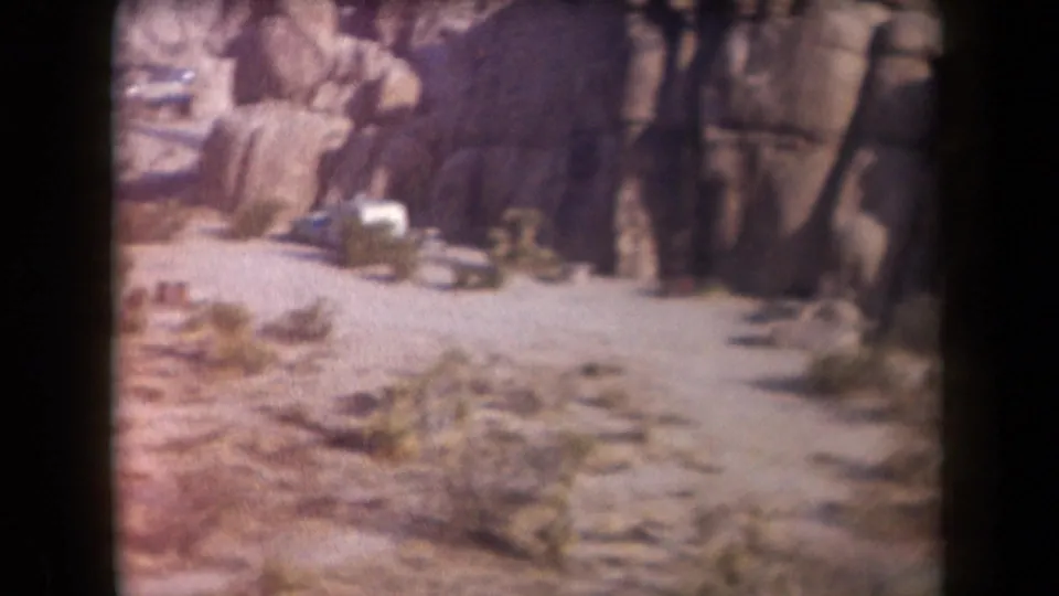 1958 Arizona desert frame showing a dirt path between rock formations and a small parked vehicle near the rocks.