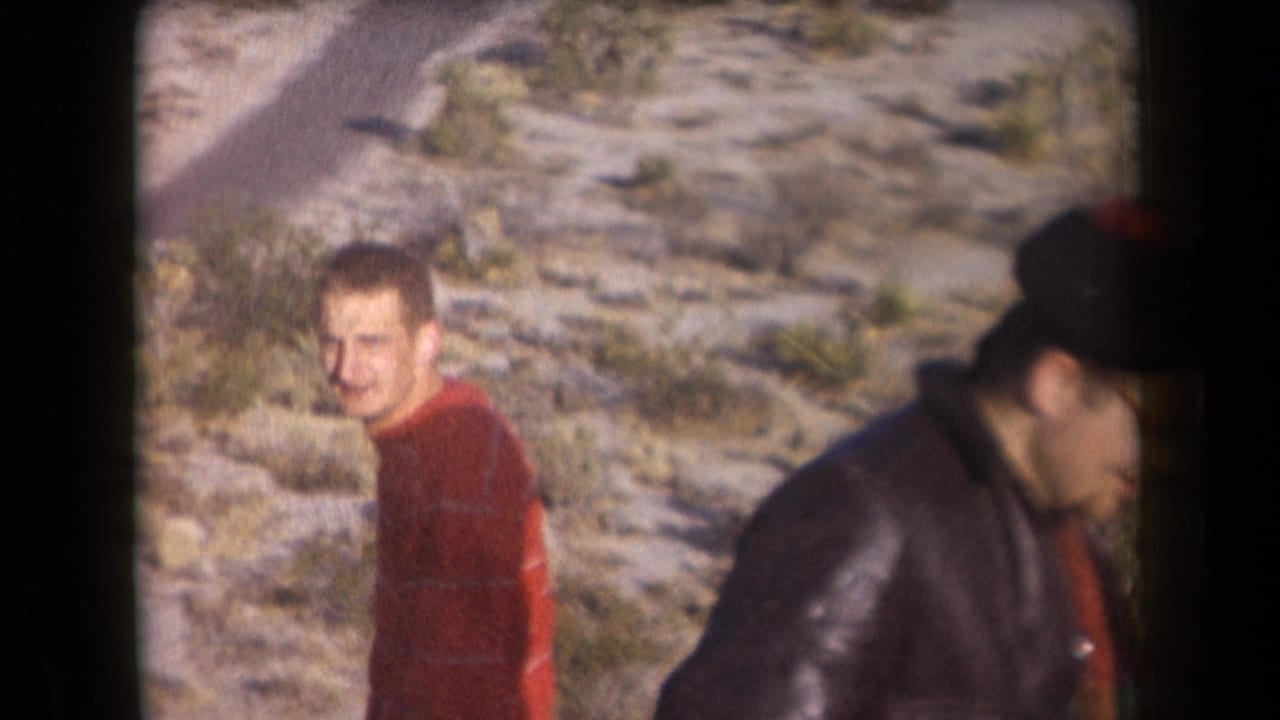 Man in a red striped sweater looks toward the camera as another man passes at right.