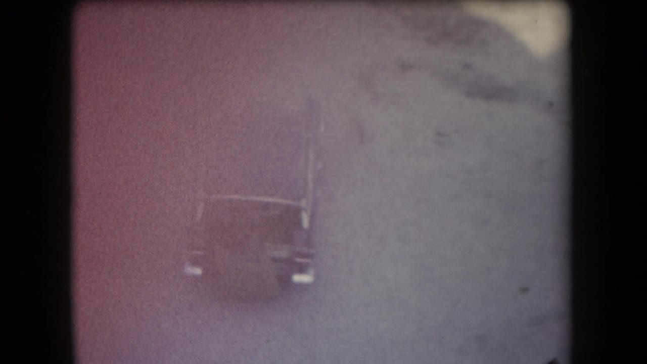 High-angle 1958 Arizona 8mm frame of a dark car on a dusty desert road, softened by faded color and grain.