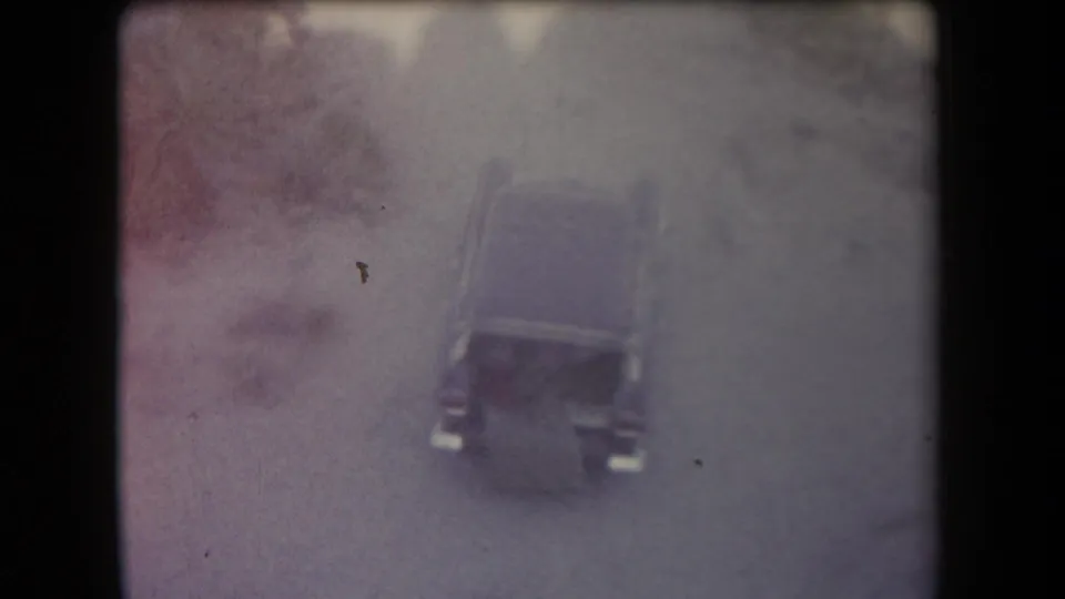 High-angle home-movie frame showing a dark car on a dusty Arizona road, with reddish brush and grainy faded color.