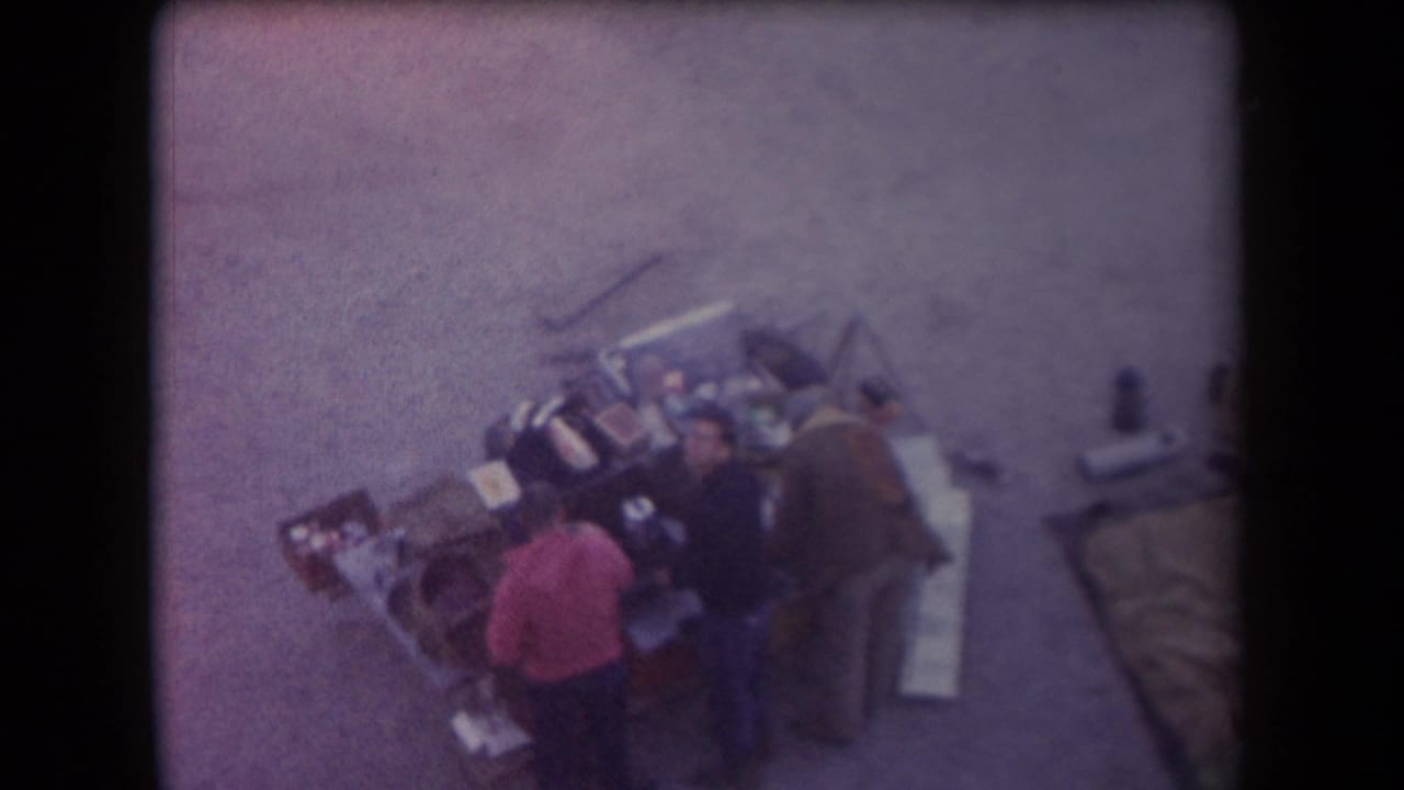 High-angle 1958 Arizona home-movie frame showing adults near an outdoor grill or serving area on paved ground.