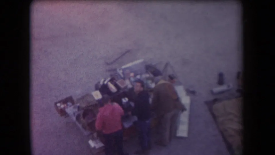 High-angle 1958 Arizona home-movie frame showing adults near an outdoor grill or serving area on paved ground.