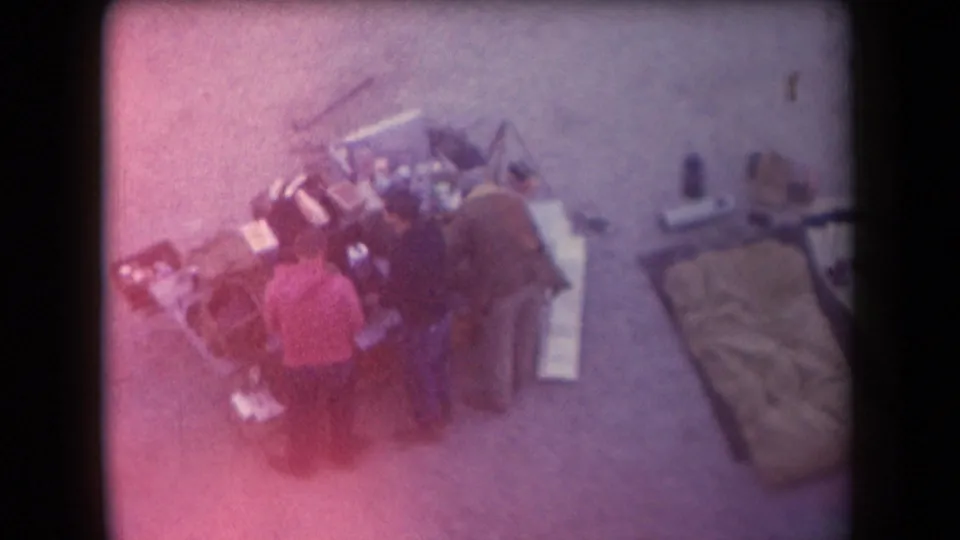 Purple-tinted 1958 Arizona 8mm frame with adults gathered by a food setup and a sleeping bag or bedroll nearby.
