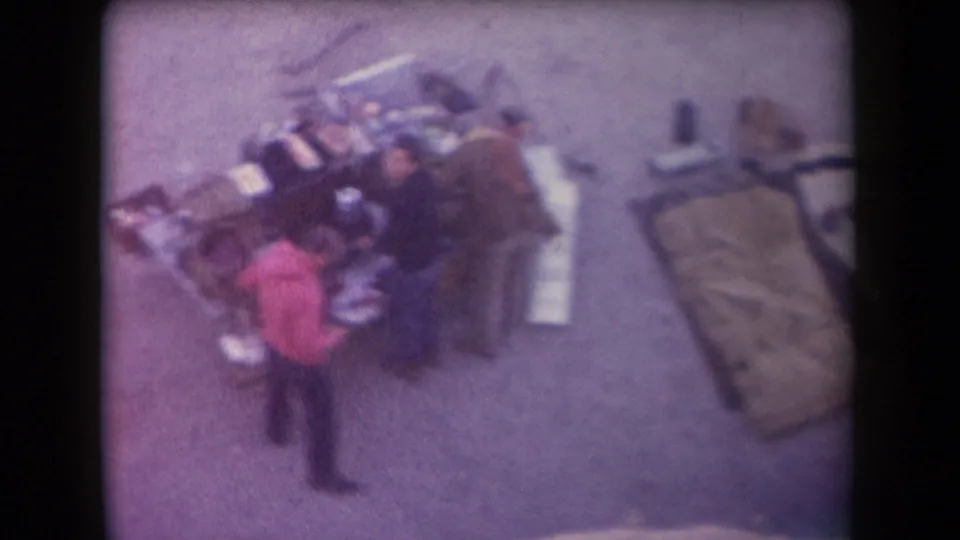High-angle 1958 Arizona home-movie frame showing adults around an outdoor grill or serving table, with a bedroll to the right.