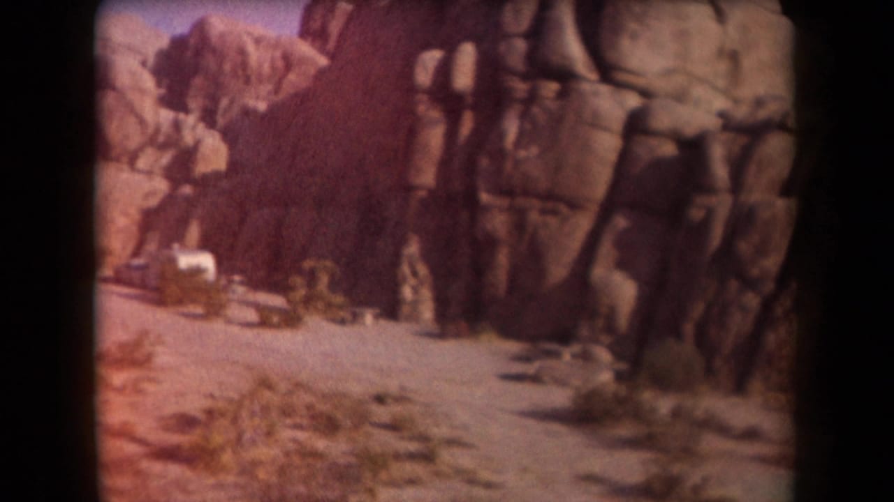 1958 Arizona home-movie frame showing a dirt road, tall weathered rocks, scrub vegetation, and a white camper trailer in the desert.