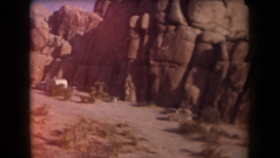 1958 Arizona home-movie frame showing a dirt road, tall weathered rocks, scrub vegetation, and a white camper trailer in the desert.