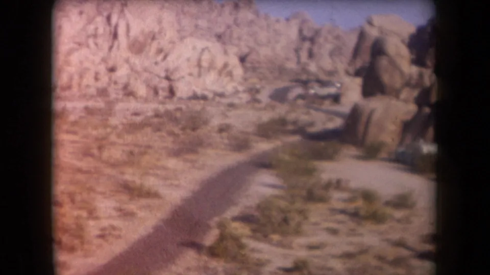 1958 Arizona desert travel frame with a curving dirt road, sparse vegetation, rocky slopes, and a distant car or trailer near the rocks.
