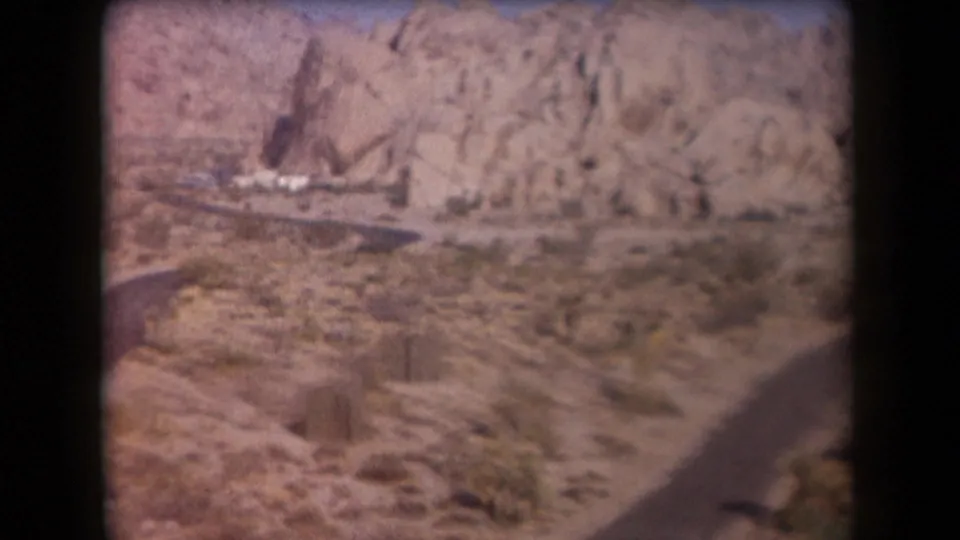 Soft 8mm Arizona desert frame showing rocky terrain, low vegetation, a dirt road edge, and a small white vehicle or trailer near the rocks.