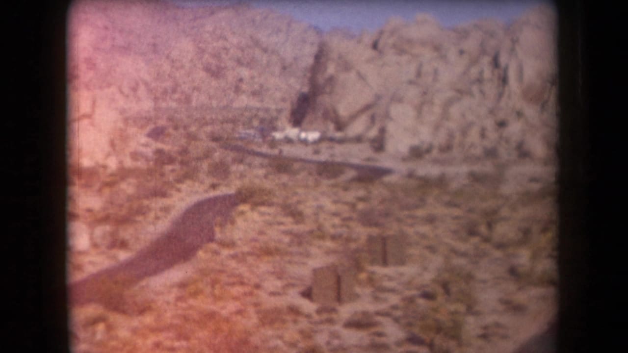 1958 Arizona 8mm frame showing a rocky desert valley, winding road, sparse scrub, and a small distant settlement.