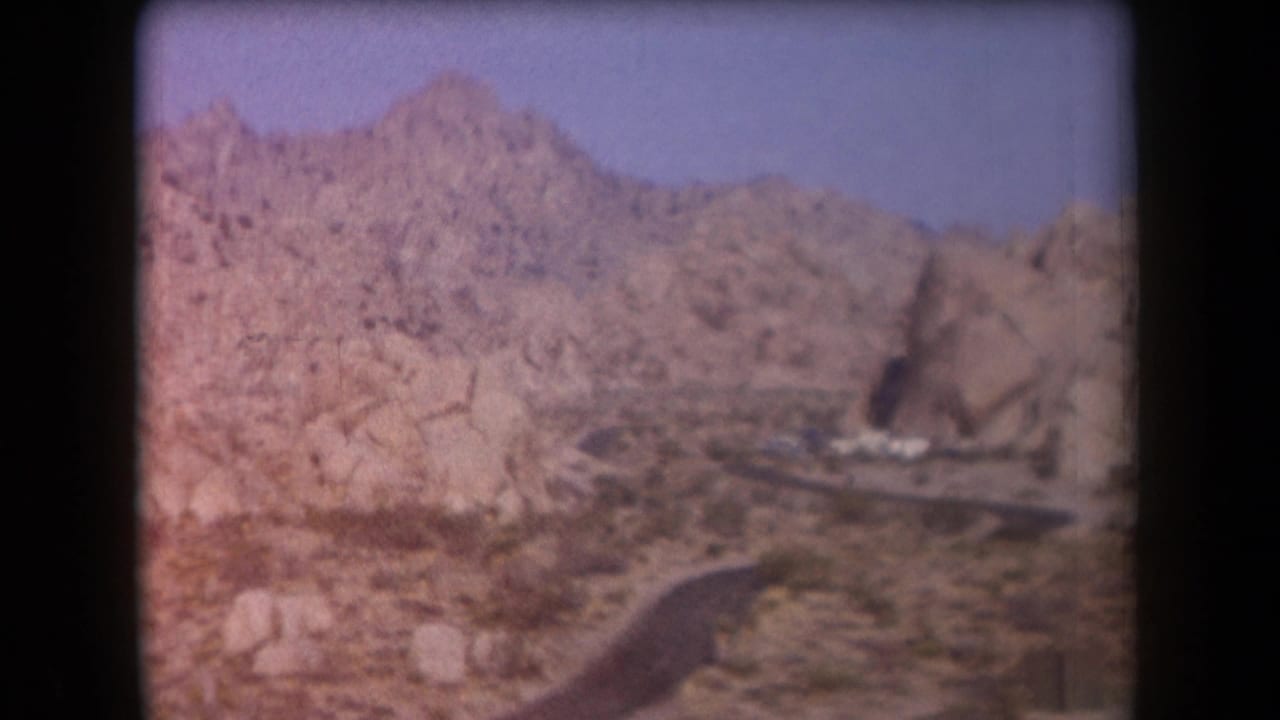 Curving desert road below rocky hills with a distant settlement.