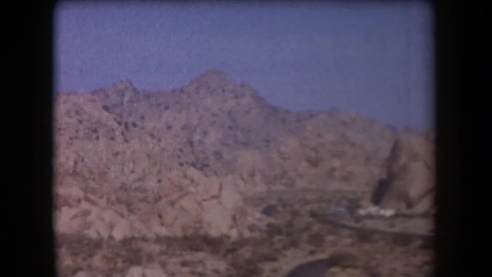 1958 Arizona 8mm frame with rugged mountain ridges, a dry valley foreground, and part of a curving road at the bottom edge.