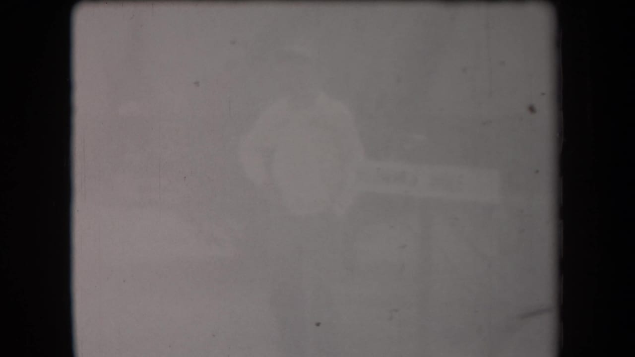 Extremely soft 1958 Arizona 8mm frame showing a pale-shirted adult figure near an indistinct informational sign-like object.