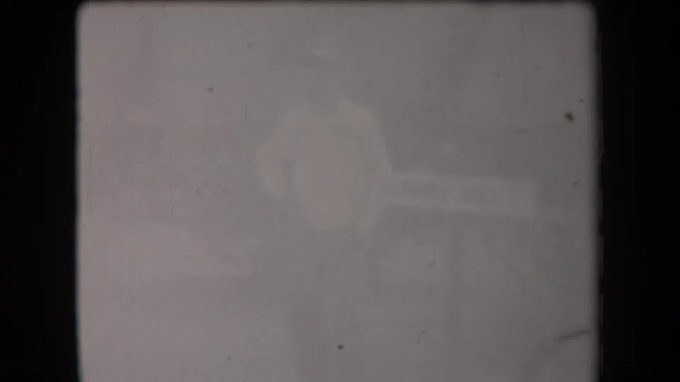 Extremely soft 1958 Arizona 8mm frame showing a pale-shirted adult figure near an indistinct informational sign-like object.