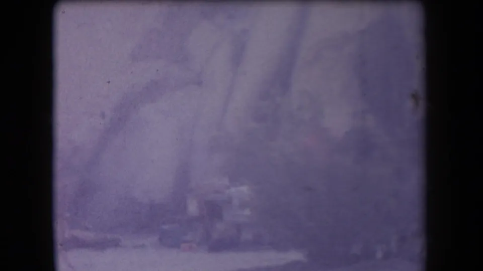 Blurry 1958 Arizona home-movie frame showing trees and two indistinct people near a small structure or table in a wooded area.