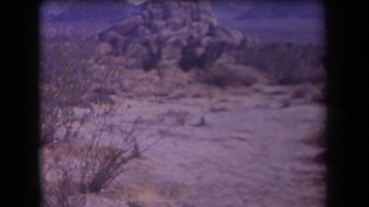1958 Arizona 8mm frame showing sandy desert ground, scrub brush, and rocky hills before the explosion.