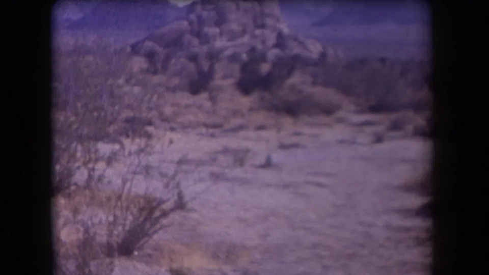 1958 Arizona 8mm frame showing sandy desert ground, scrub brush, and rocky hills before the explosion.