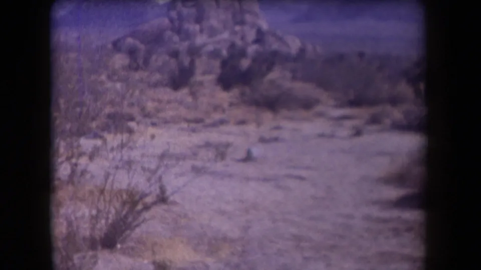 1958 Arizona desert view on aged 8mm film, with scrub, pale ground, and rocky hills visible before smoke appears.