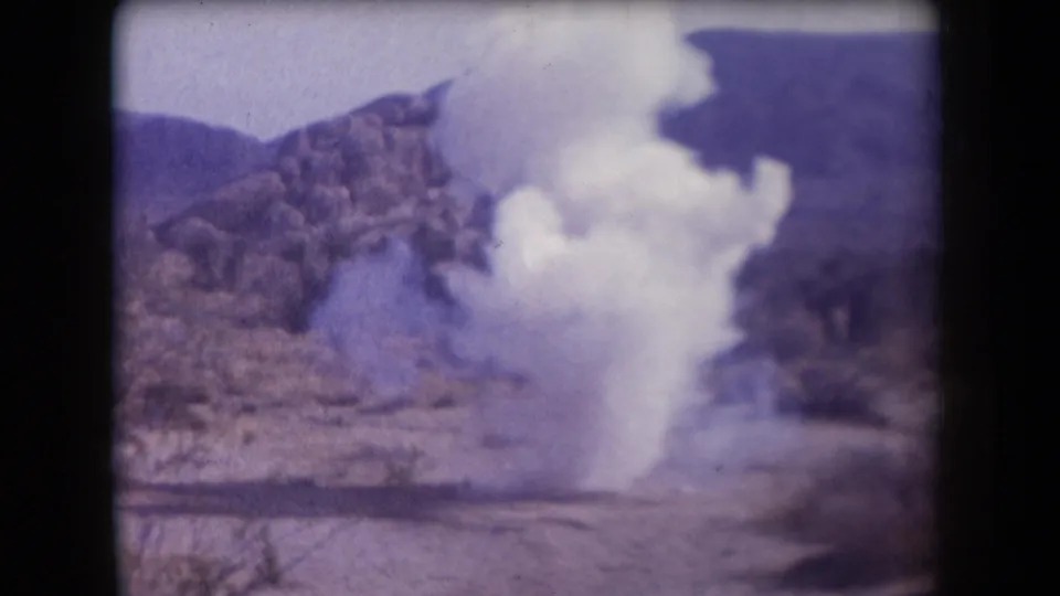 1958 Arizona 8mm frame showing a pale smoke or dust plume rising from desert ground below rocky hills.