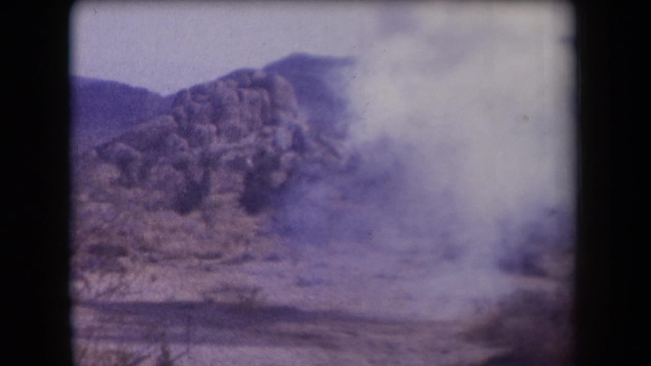 1958 Arizona 8mm frame with a spreading smoke or dust cloud partly obscuring desert rocks and low mountains.