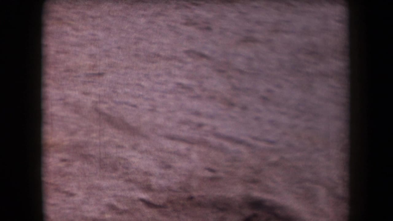 1958 Arizona desert ground appears in a soft handheld 8mm frame with reddish sand, small ridges, stones, and dark gate edges.