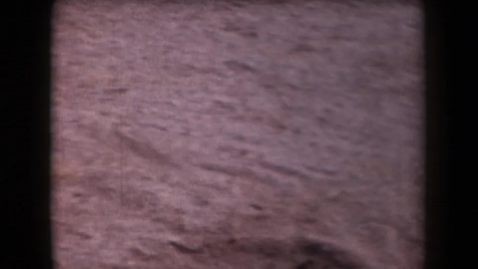 1958 Arizona desert ground appears in a soft handheld 8mm frame with reddish sand, small ridges, stones, and dark gate edges.