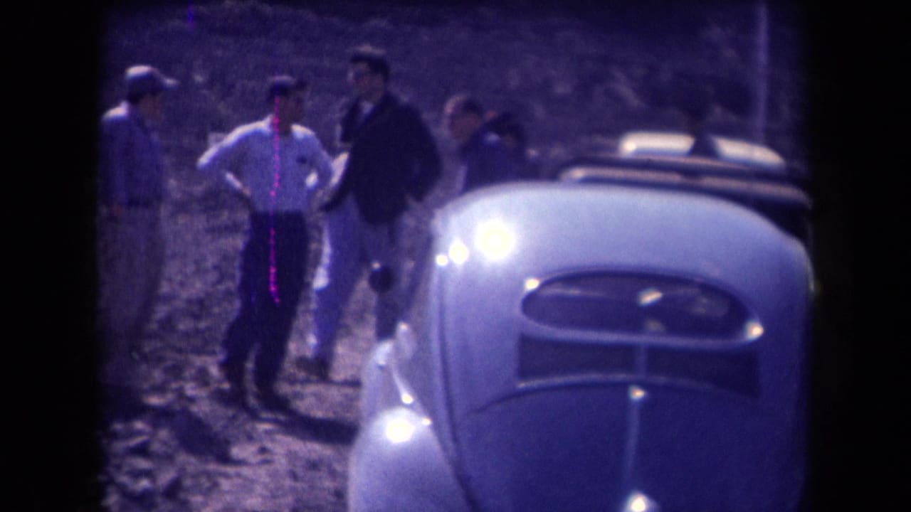 Several adult men stand beside a light-colored Beetle on a dusty roadside.