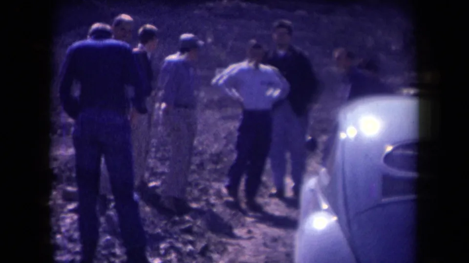 1958 Arizona home-movie frame showing men standing and walking near the front of a light-colored Beetle on uneven dirt.