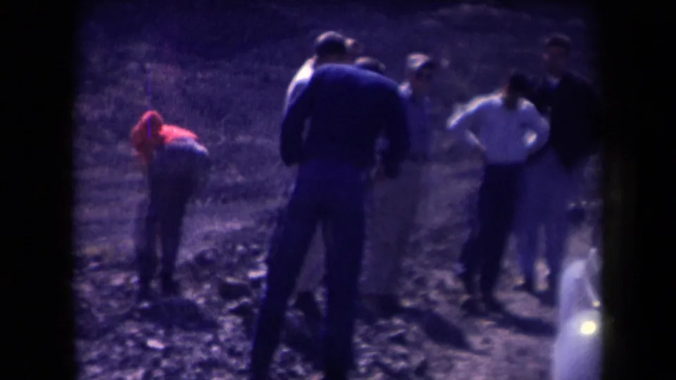 1958 Arizona frame with adult men near a light-colored Beetle as one person in red bends down on the dusty roadside.