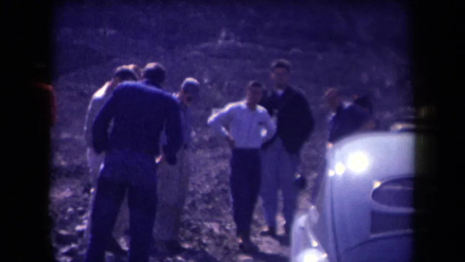 In 1958 Arizona footage, several men gather on a dusty roadside near a light-colored mid-century car.