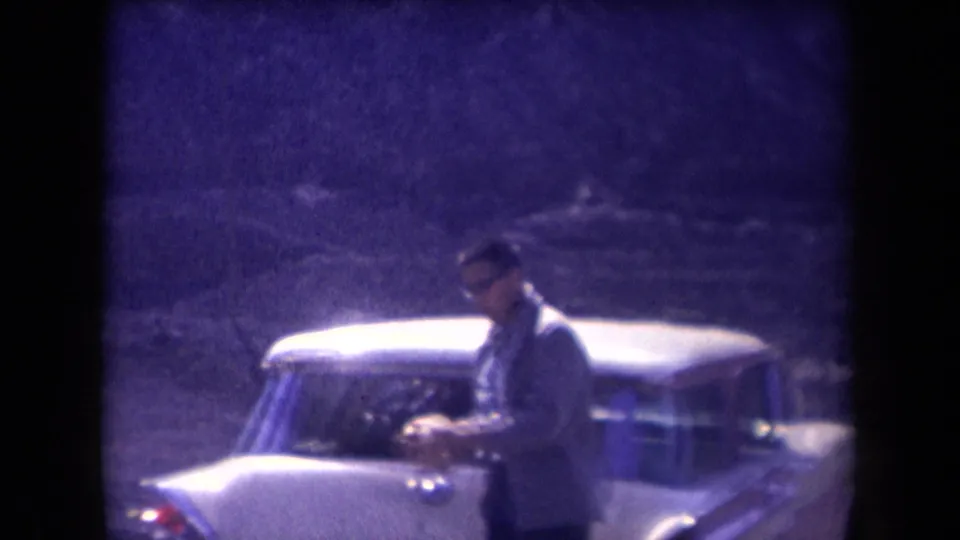 A man in mid-century clothing stands by a light-colored sedan in the Arizona roadside home movie.
