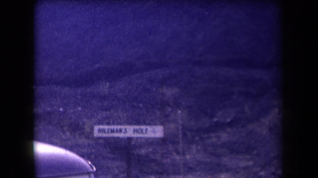 1958 Arizona home-movie frame showing a purple-tinted hillside, a sign reading Mileman's Hole, and a partial car roof in the foreground.