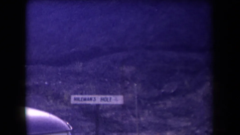1958 Arizona home-movie frame showing a purple-tinted hillside, a sign reading Mileman's Hole, and a partial car roof in the foreground.