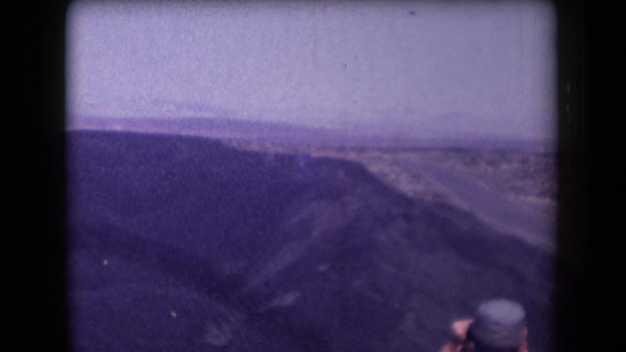 1958 Arizona home-movie frame showing a canyon overlook, winding highway, pale sky, and a partially seen traveler at the bottom.