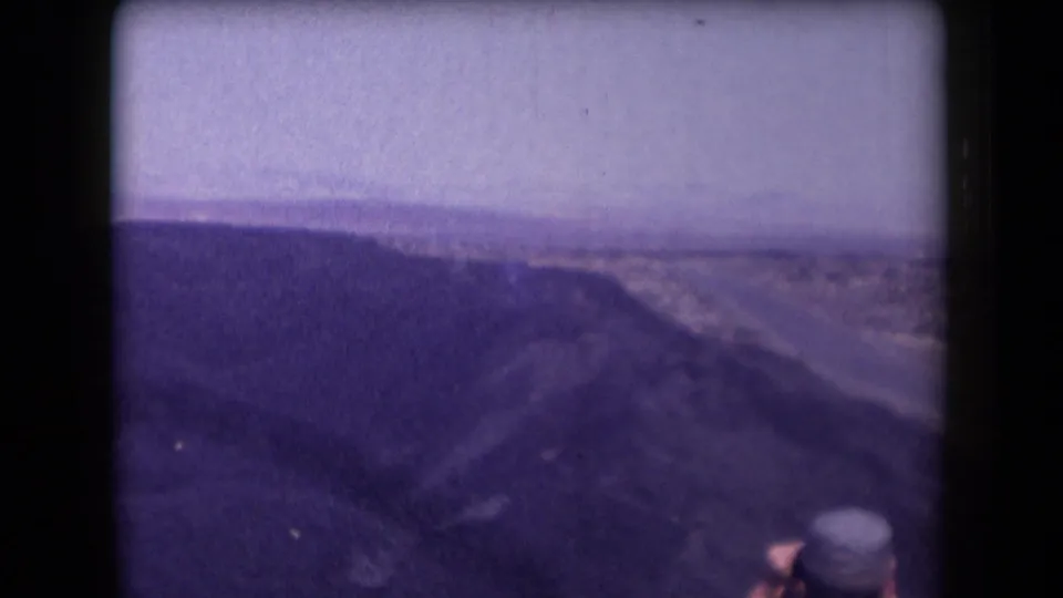 1958 Arizona home-movie frame showing a canyon overlook, winding highway, pale sky, and a partially seen traveler at the bottom.