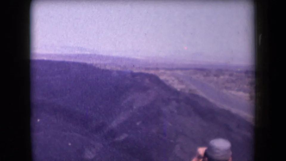 Arizona home-movie frame from 1958 showing a canyon slope, highway, distant desert, and a cropped traveler at the lower right.
