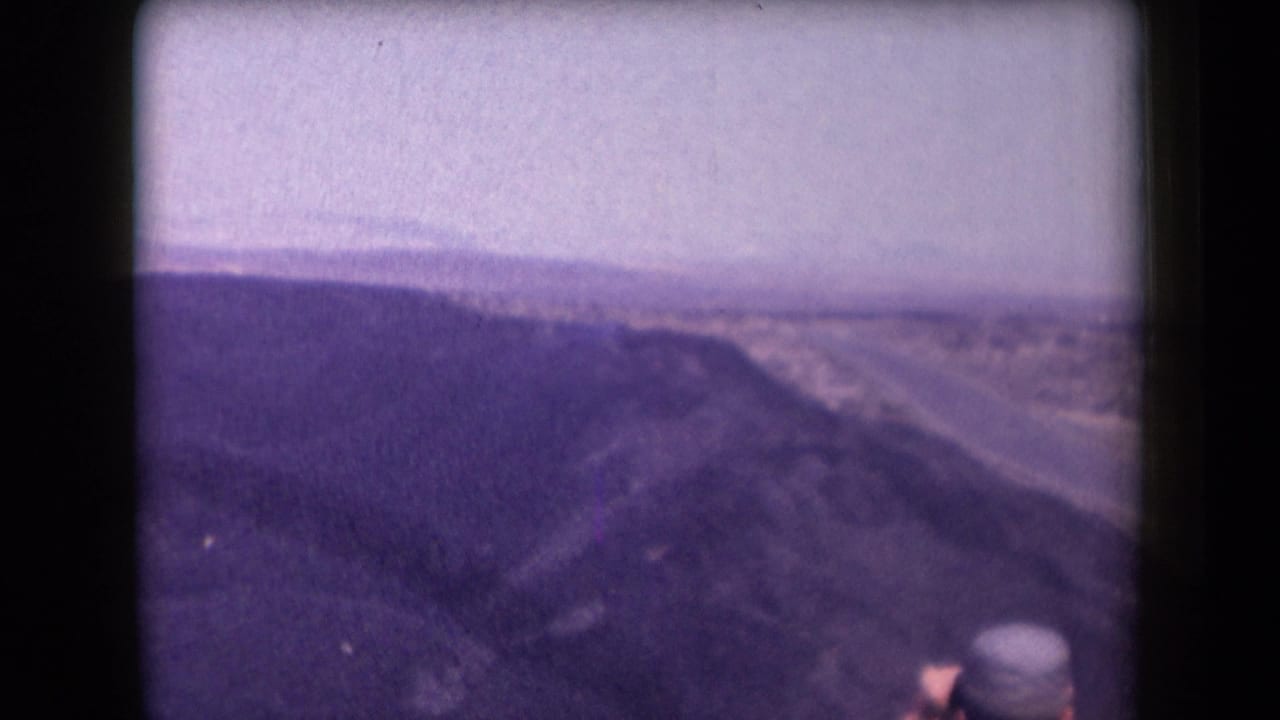 1958 Arizona 8mm frame showing a desert ridge, hazy horizon, and a partially visible person in a hat near the lower foreground.