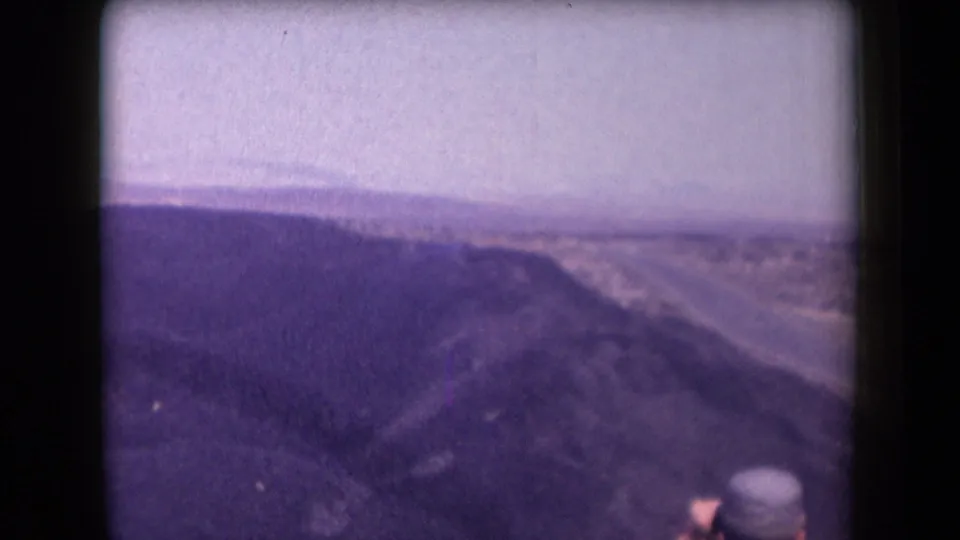1958 Arizona 8mm frame showing a desert ridge, hazy horizon, and a partially visible person in a hat near the lower foreground.