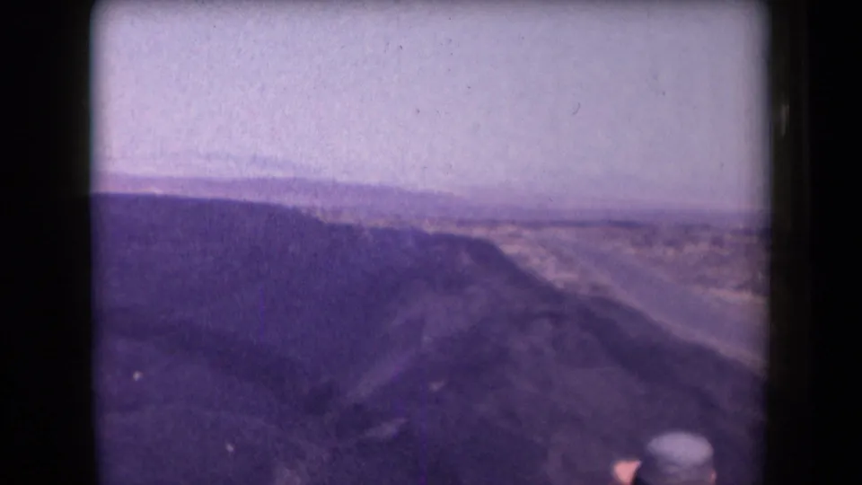 1958 Arizona home-movie frame with open desert terrain, distant horizon, and a partly visible hat-wearing observer at the lower right.