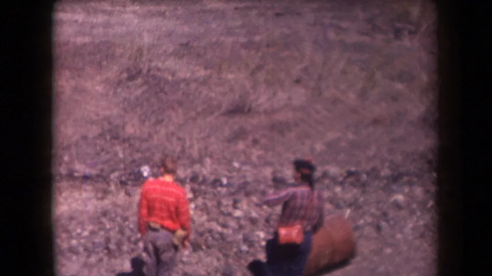 Two hikers, including one in a red sweater, walk uphill across rocky Arizona terrain in a 1958 8mm home movie.