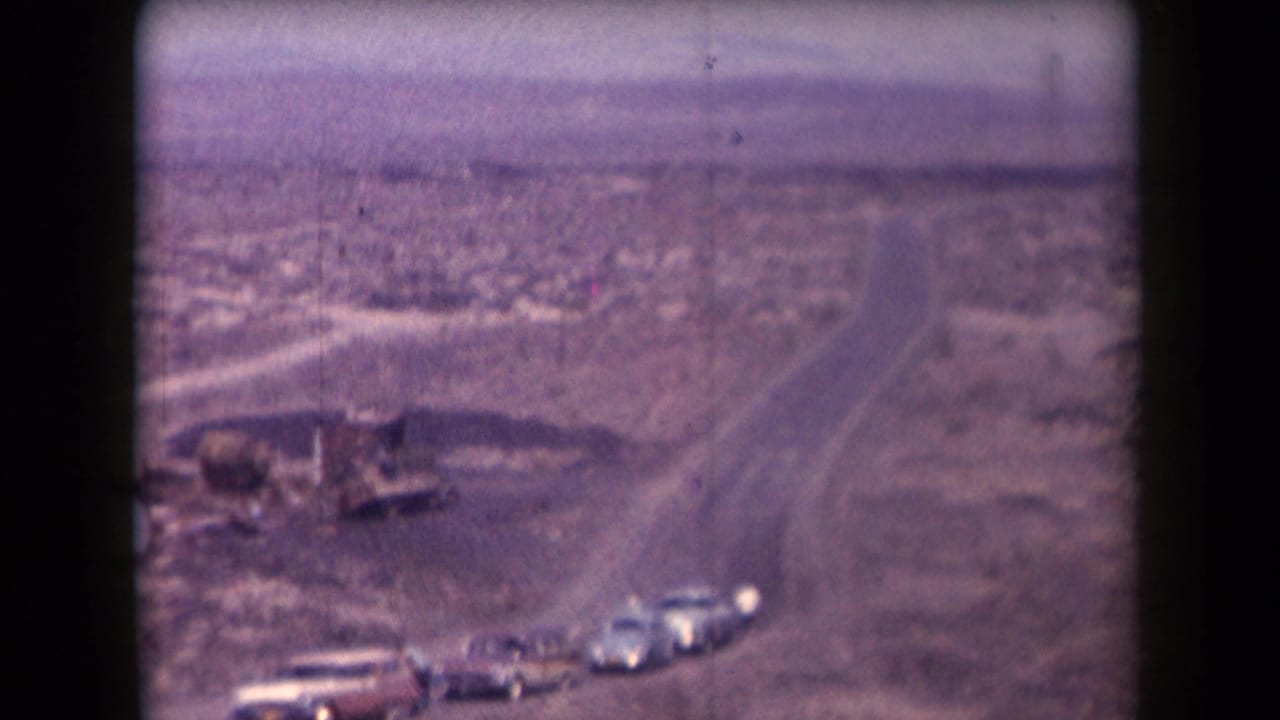 1958 Arizona home-movie frame showing cars on a desert highway with sparse roadside structures and distant open land.