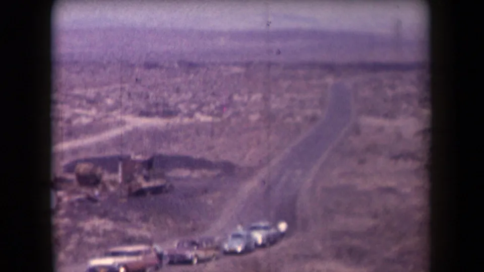 1958 Arizona home-movie frame showing cars on a desert highway with sparse roadside structures and distant open land.