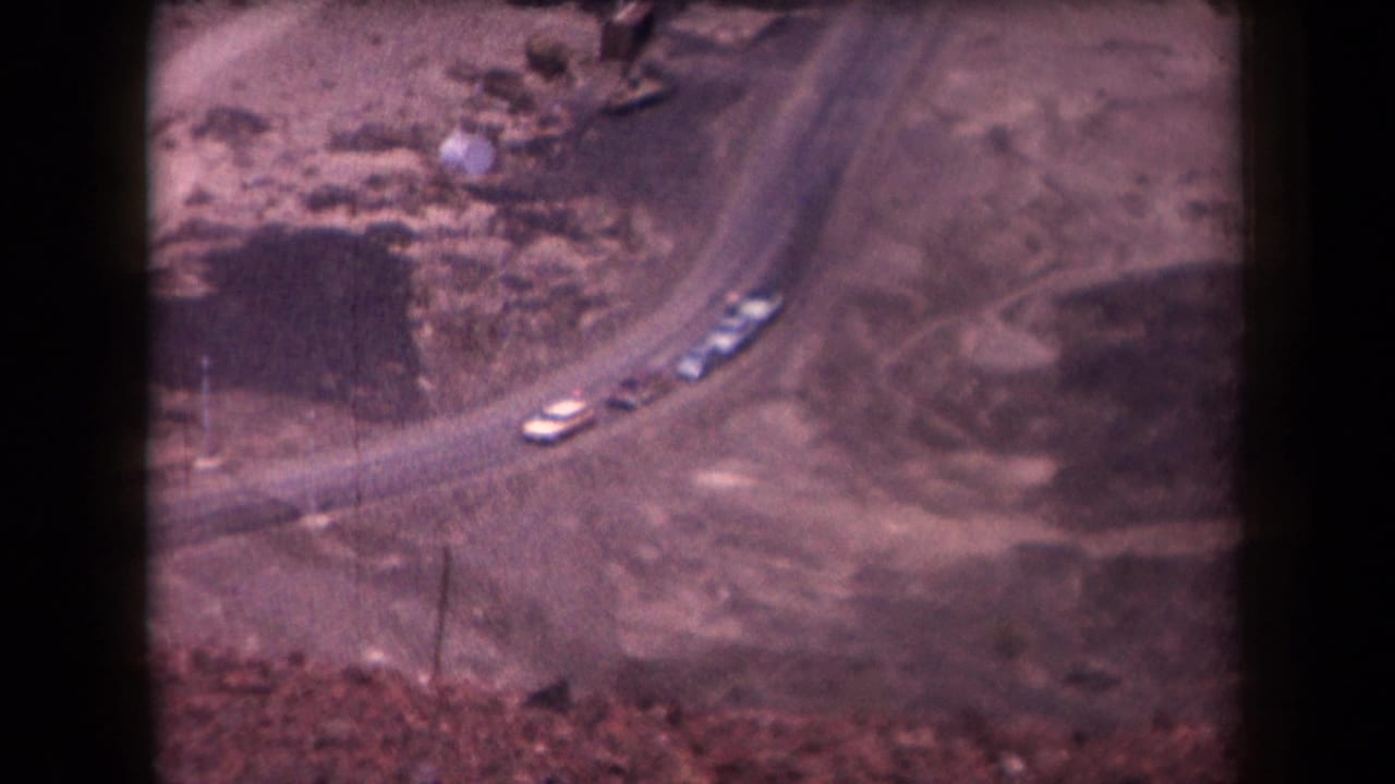 Cars continue along a curved dirt road seen from above.