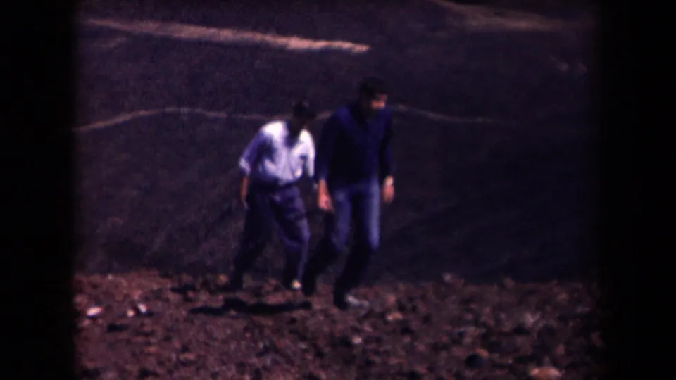 1958 Arizona home-movie frame showing two people walking uphill over rocky desert ground near a road edge.