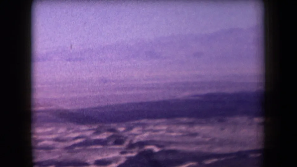 1958 Arizona desert footage frame with pale sandy flats, distant low hills, hazy sky, and visible 8mm gate edges.