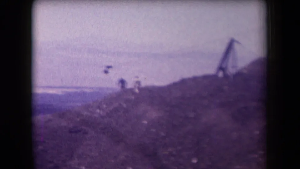 1958 Arizona 8mm frame showing distant people on a rocky desert hill near a simple zipline structure under a hazy sky.