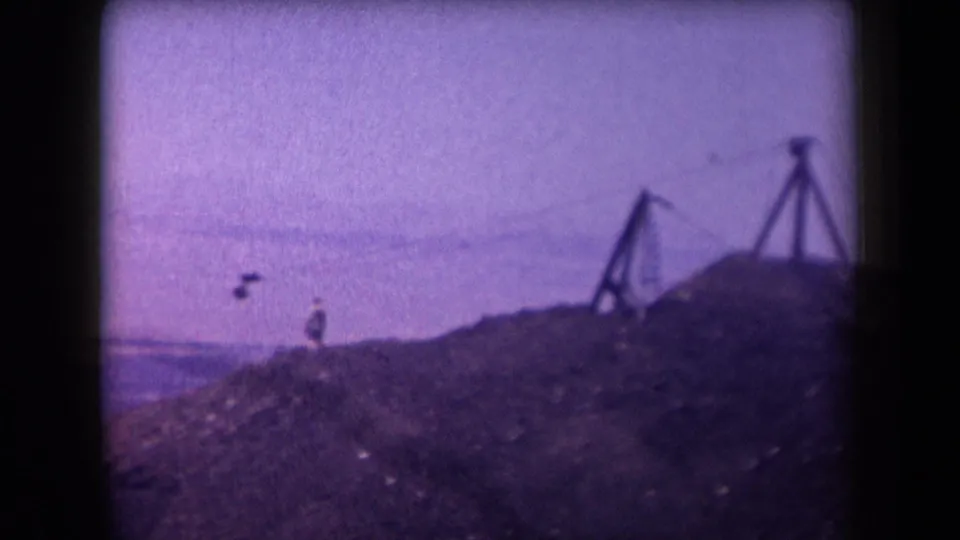 Purple-tinted 1958 Arizona home-movie frame with a small rider on a zipline beside rough desert ground and wooden support frames.