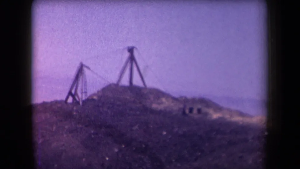 1958 Arizona 8mm frame focused on simple wooden zipline supports on a rocky hill, with distant arid terrain behind them.