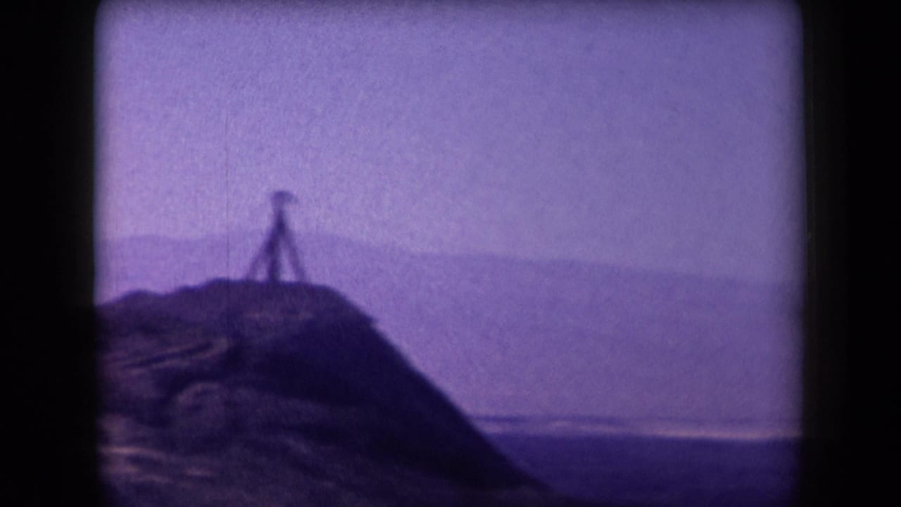 1958 Arizona 8mm frame showing a hazy hilltop, distant mountains, and a small dark silhouette that may be a person or tripod on the ridge.