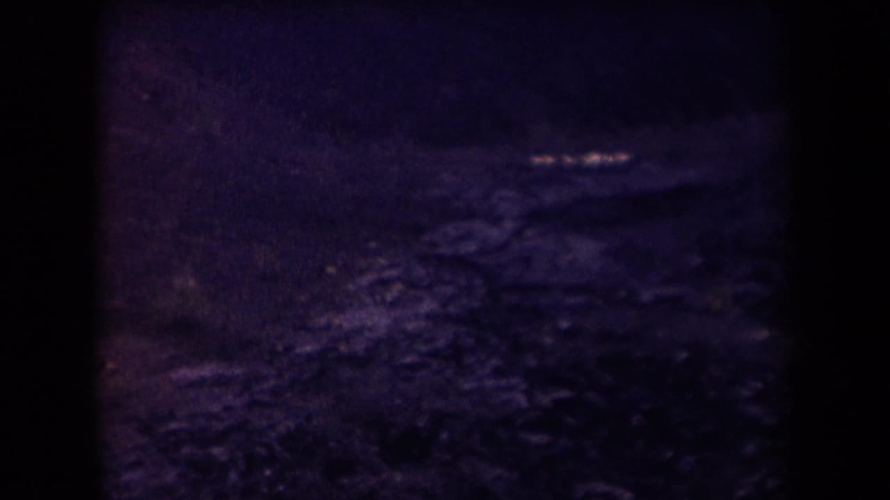 1958 Arizona 8mm frame showing a dark, purple-tinted rocky landscape below an aircraft window, with heavy shadow and soft grain.