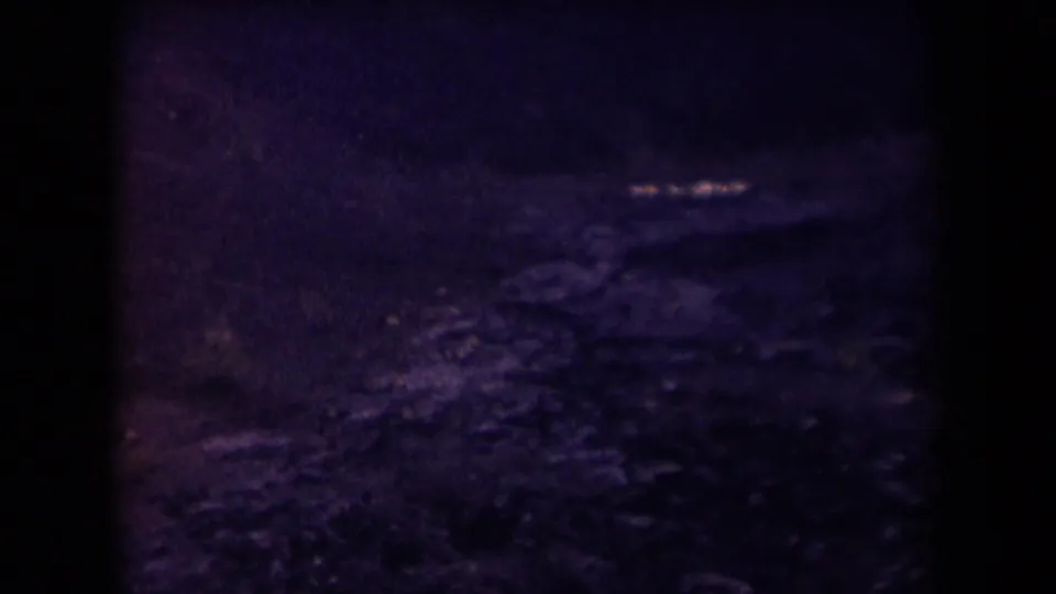 1958 Arizona 8mm frame showing a dark, purple-tinted rocky landscape below an aircraft window, with heavy shadow and soft grain.